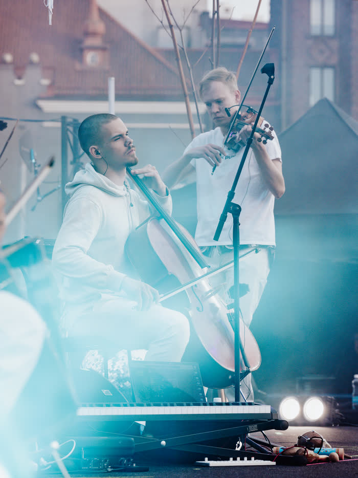 Kaur Nellis playing cello in a white hoodie with a keyboard and laptop set up in front of him, a Nordic Pulse violinist behind, washed in pale blue light.