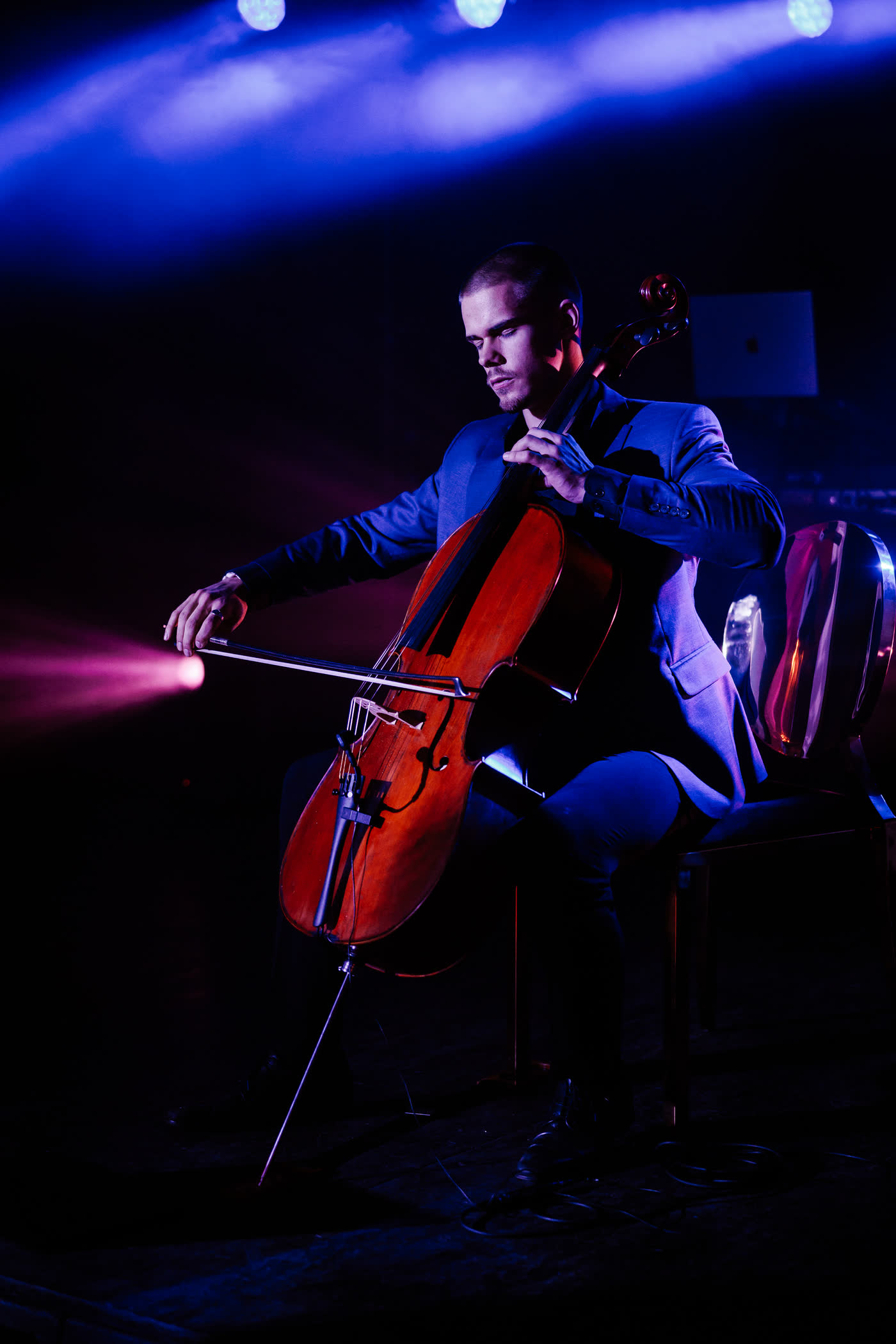 Kaur Nellis playing cello alone on a darkened stage, lit deep blue with a magenta beam cutting across his bow.